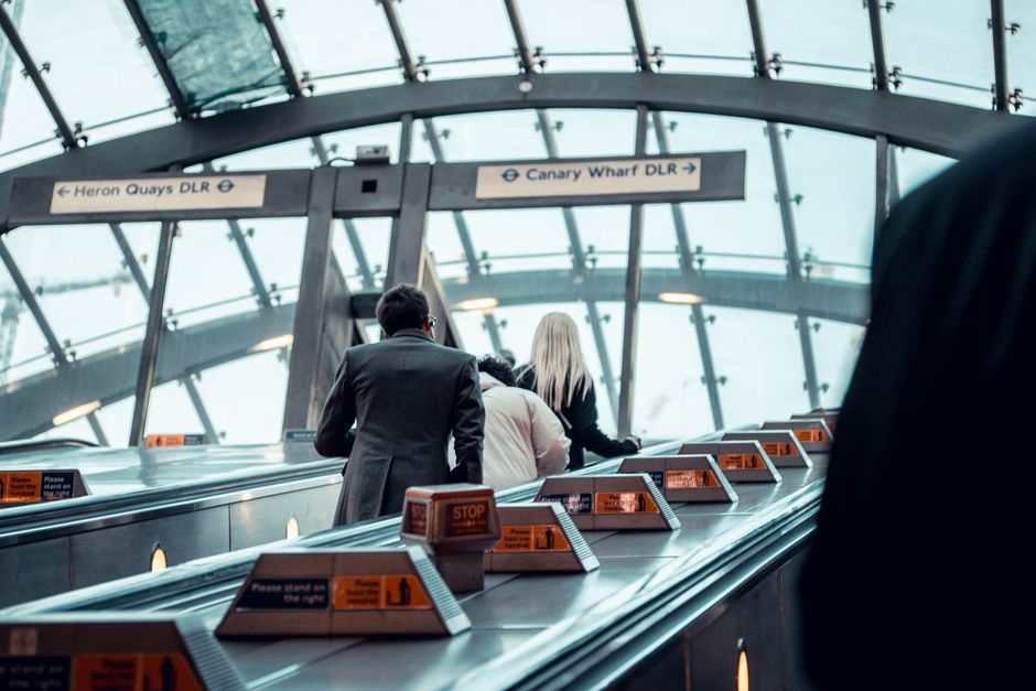Inside a modern transportation terminal with a glass-paneled curved ceiling allowing natural light to fill the space, two people are standing at a metal luggage conveyor belt, which is lined with oran