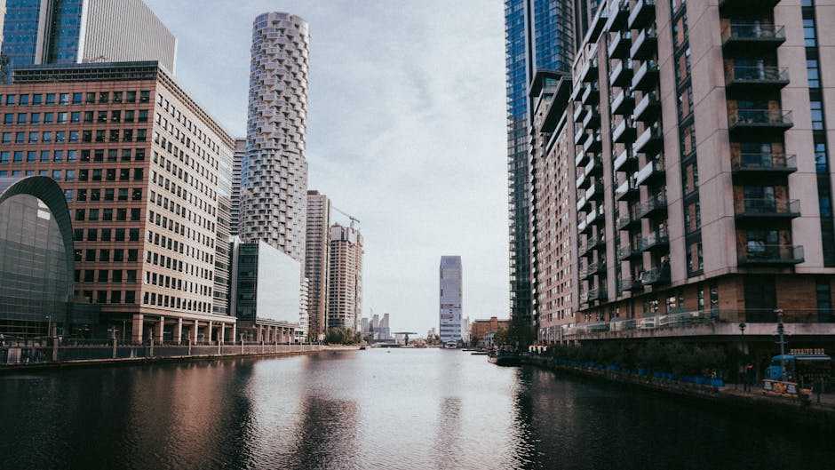 A cityscape featuring a modern waterfront area with several high-rise office buildings crafted from glass and steel, reflecting the blue sky and surrounding structures. The foreground shows a black me