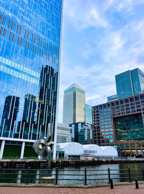 A nighttime cityscape of Canary Wharf featuring illuminated skyscrapers with modern glass and steel facades, reflecting colorful lights onto the calm water of the river in the foreground. The building