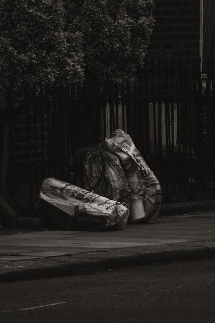 A city street scene featuring a white waste collection truck with an open rear compactor area, showing signs of rust and used for rubbish collection. The truck is parked parallel to a row of multi-sto